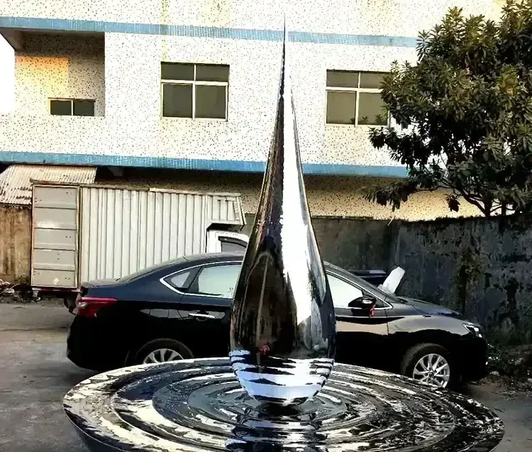 Polished stainless steel water droplet sculpture above a rippled metal fountain base in an outdoor yard