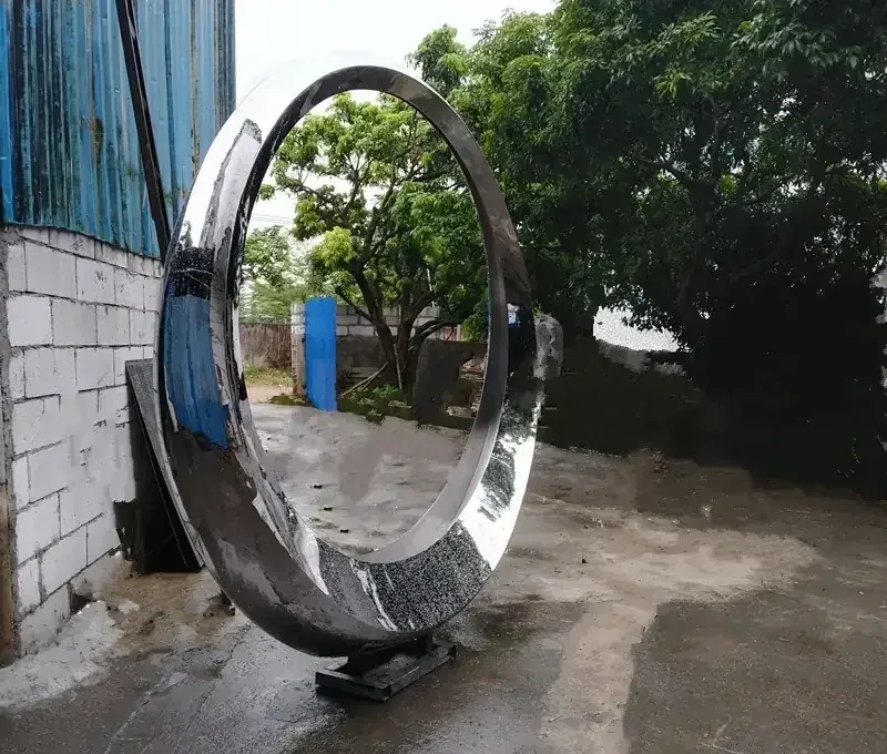 Large mirror-polished stainless steel ring sculpture standing in an outdoor workshop yard beside a white brick wall