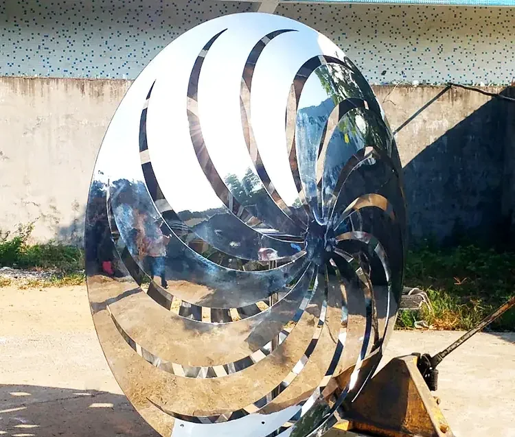 Large mirror-polished stainless steel disc sculpture with spiral swirl cutouts standing outdoors against a concrete wall