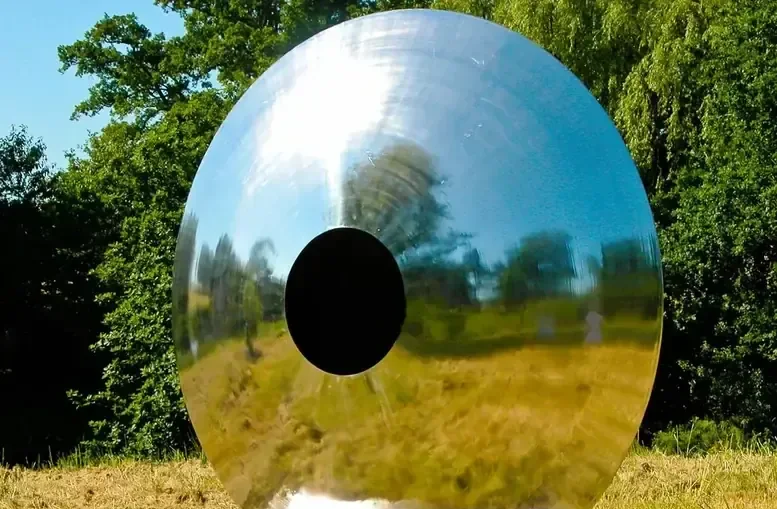A large, highly polished metal disc with a central hole stands in a grassy field.