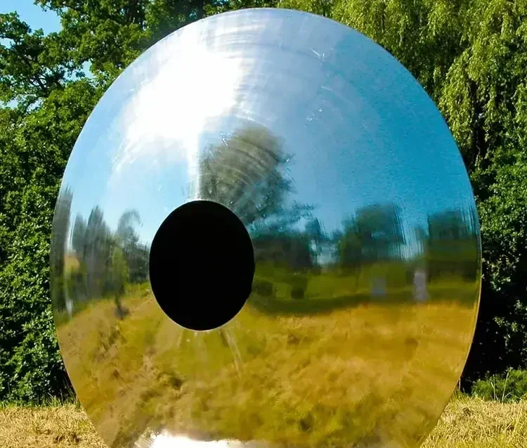 A large, circular, reflective metal disc with a central hole, standing outdoors.