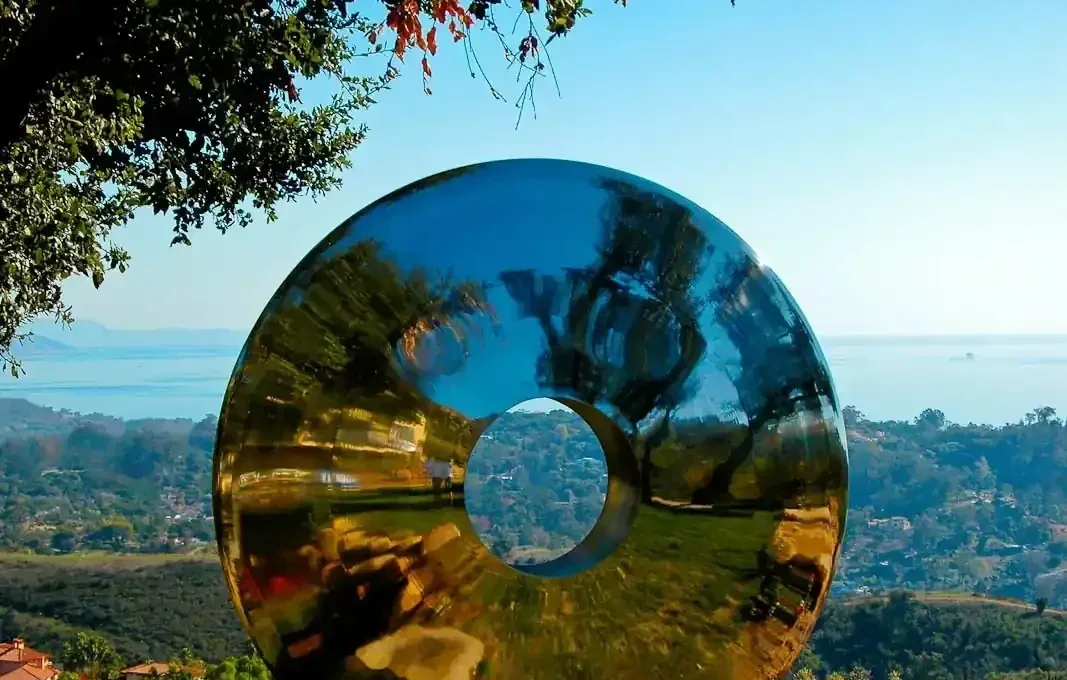 Large polished metal torus sculpture outdoors reflecting trees, hills, and ocean under a clear blue sky