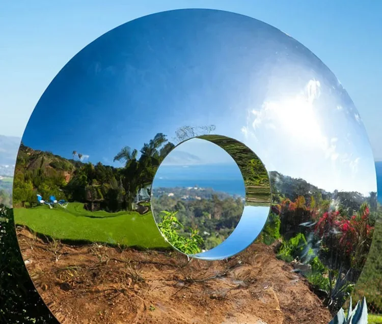 Mirror-polished stainless steel torus sculpture outdoors reflecting blue sky, clouds, trees and a coastal hillside