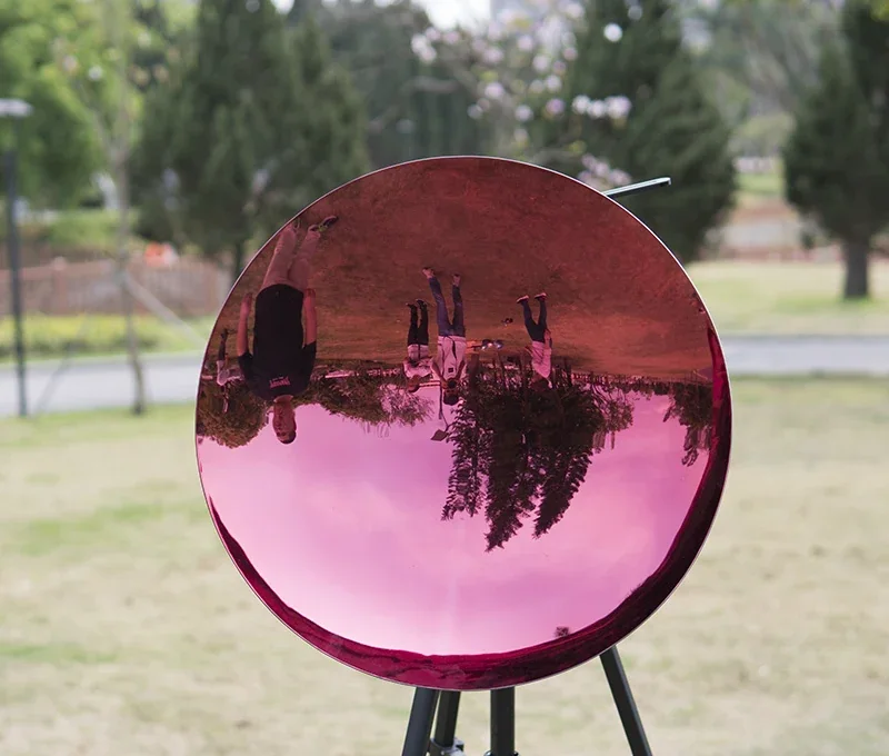 A large, pink, concave mirror on a tripod outdoors, reflecting an inverted view of people and trees.