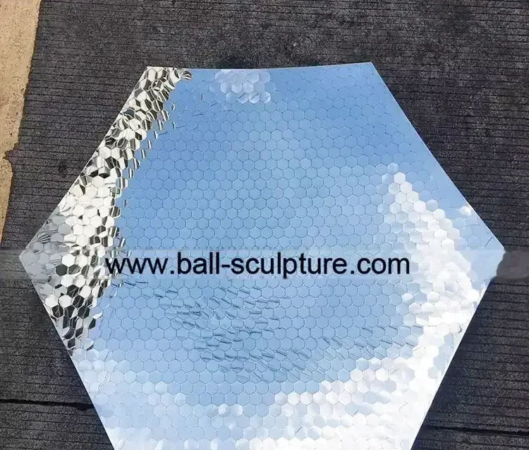 Hexagonal panel covered in small mirror tiles reflecting sky, lying on dark pavement with website URL overlaid
