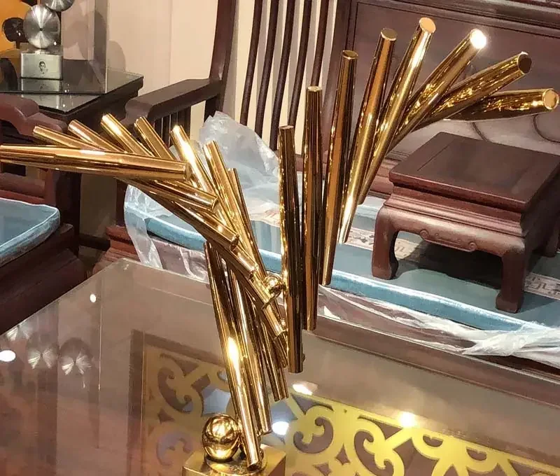 Gold-tone metal sculpture of angled tubes arranged in a fan shape on a glass table, with wooden furniture and plastic wrap behind
