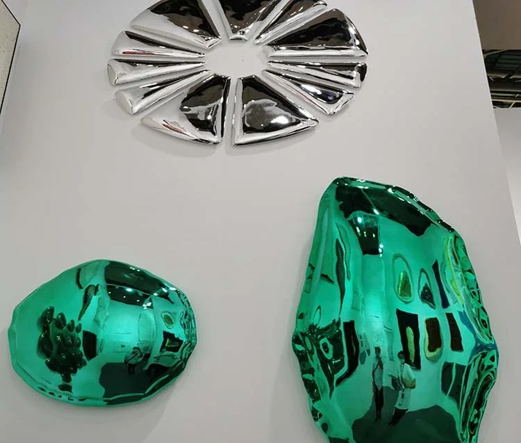 Three metallic sculptures, one silver and two green, with reflective surfaces arranged on a white display surface.