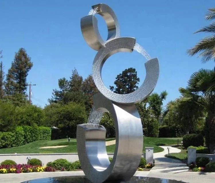 An abstract stainless steel fountain sculpture with flowing water in a landscaped garden.