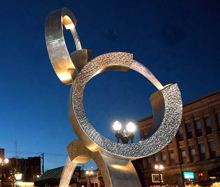 Large abstract metal ring sculpture with flowing water in a city square at dusk, street lamps and brick buildings behind