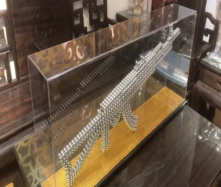 Clear acrylic display case containing a rifle-shaped sculpture made of silver magnetic balls on a gold base