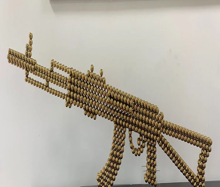 Rifle-shaped sculpture built from small golden magnetic spheres against a plain white wall