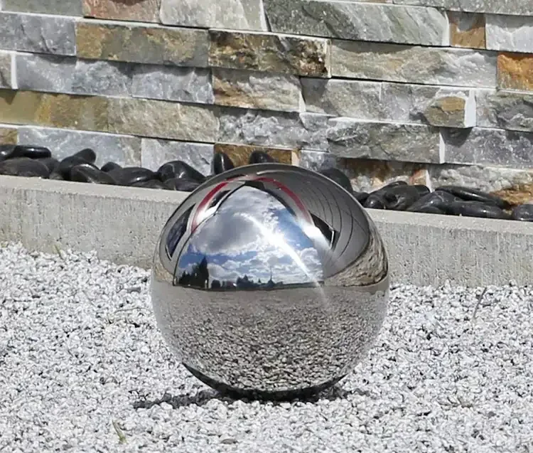Polished stainless steel sphere resting on gravel in front of a stacked slate stone wall
