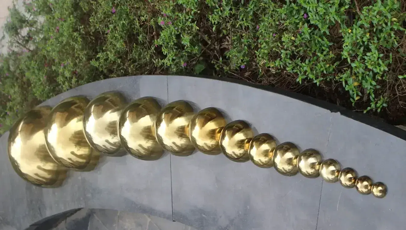 Polished golden metal spheres displayed in descending sizes on a curved stone ledge with green shrubs behind