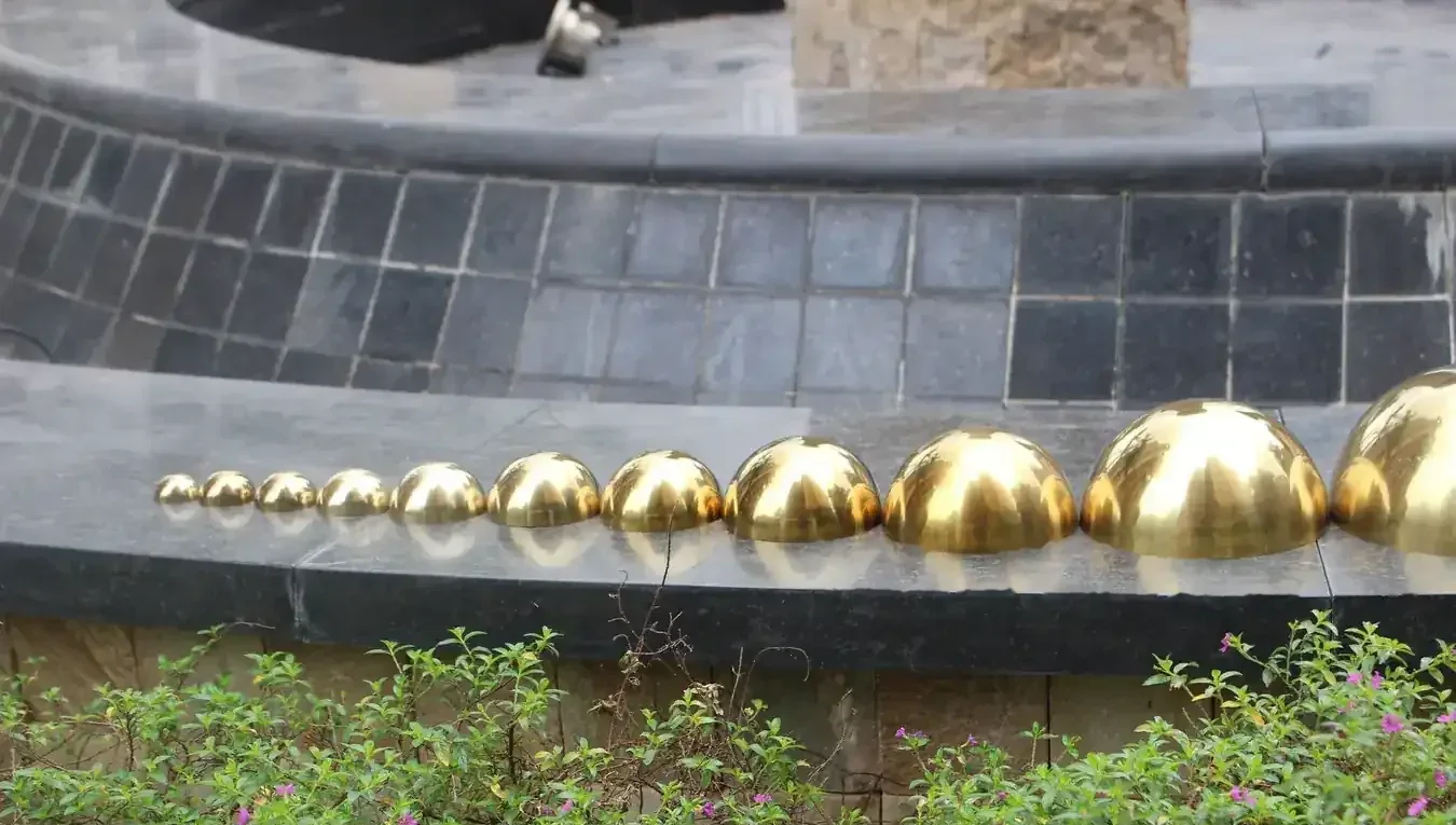 Row of polished golden brass dome halves arranged by increasing size on an outdoor ledge with tiled wall and greenery