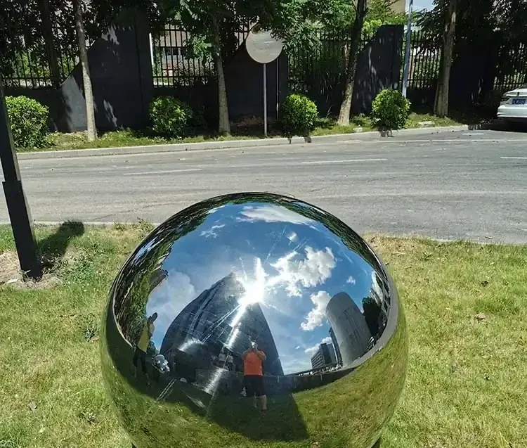 Large polished stainless steel sphere displayed on grass beside a road, reflecting trees, sky, and surroundings