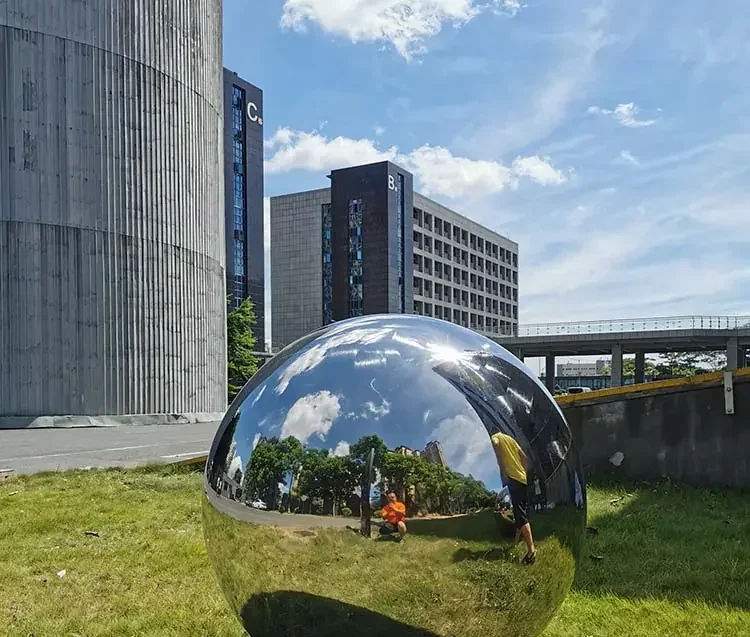 Large reflective metal sphere sculpture on a lawn in front of modern office buildings.