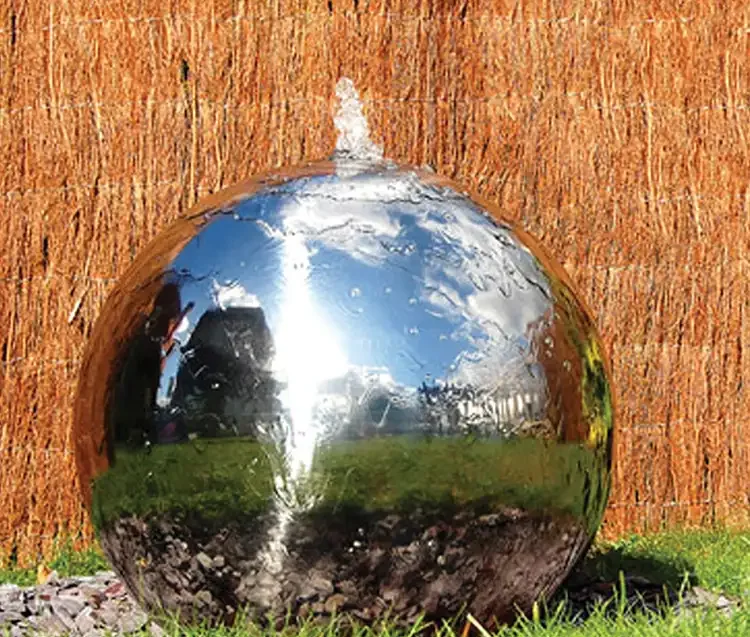 Polished stainless steel sphere water fountain spraying water from its top, set on pebbles and grass with a reed fence behind