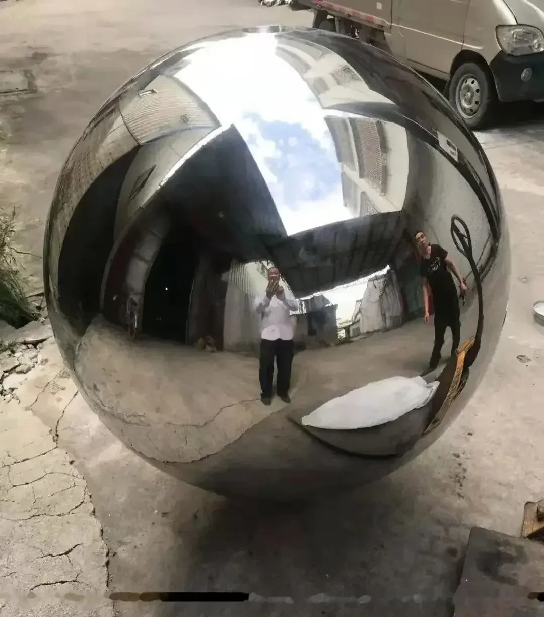 Large mirror-polished stainless steel sphere outdoors on cracked concrete, reflecting people, a truck and buildings