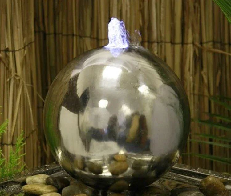 Polished stainless steel sphere water fountain spraying water, set on pebbles with bamboo fence behind