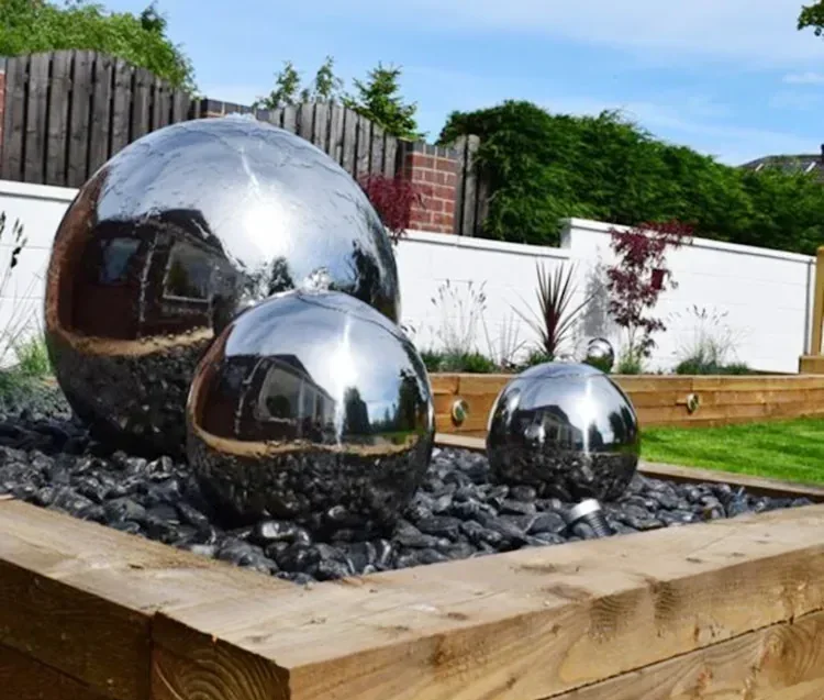 Three polished stainless steel spheres of varying sizes placed on a bed of black gravel in a raised wooden planter