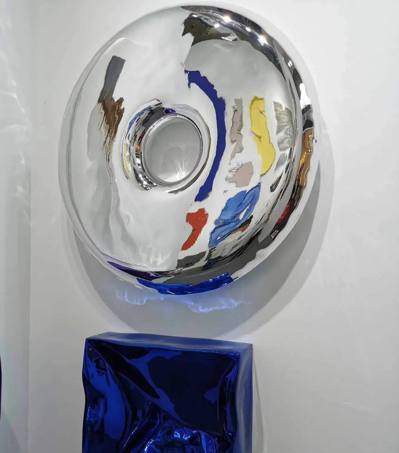 Polished mirror-finish circular wall sculpture on a white wall above a reflective blue crumpled object