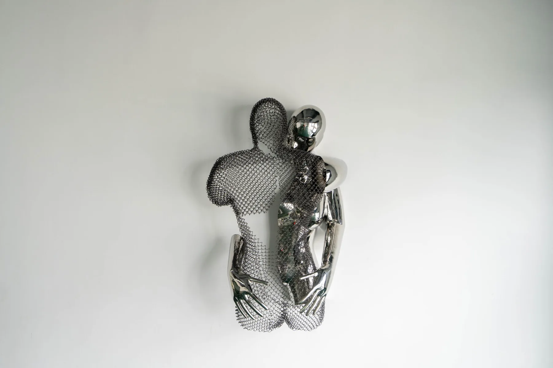 Chrome and wire mesh metal sculpture of two embracing human figures mounted on a plain white wall