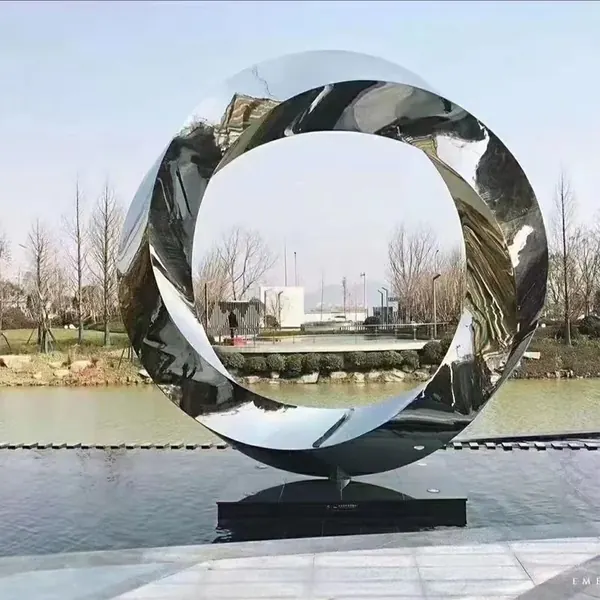 Large, reflective, stainless steel torus sculpture standing on a platform in a body of water.