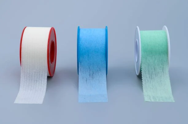 Three rolls of medical tape in white, blue, and green on plastic spools against a plain studio background