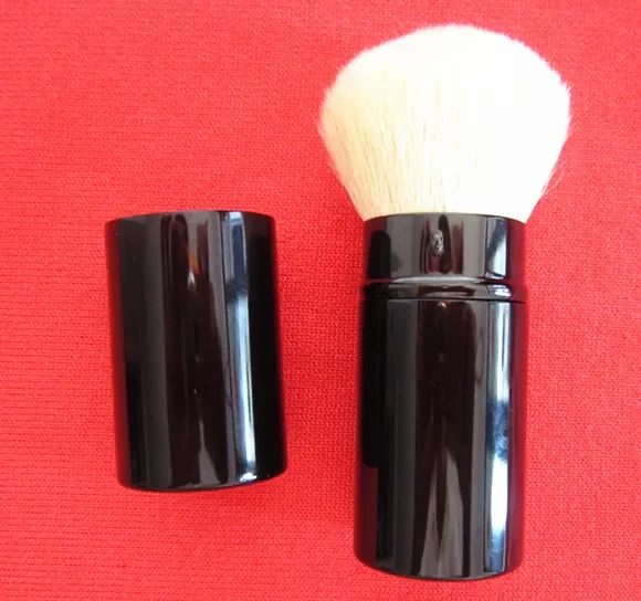 Bronze retractable powder brush with white goat hair and a shiny black tube shown on a red background