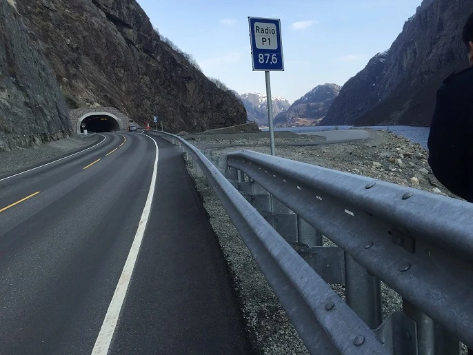 A paved road curves toward a tunnel entrance through a rocky fjord landscape, with a radio sign visible.
