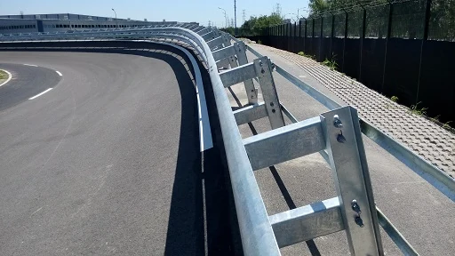 Galvanized steel W-beam guardrail along a curved asphalt road with cobblestone walkway and metal fence