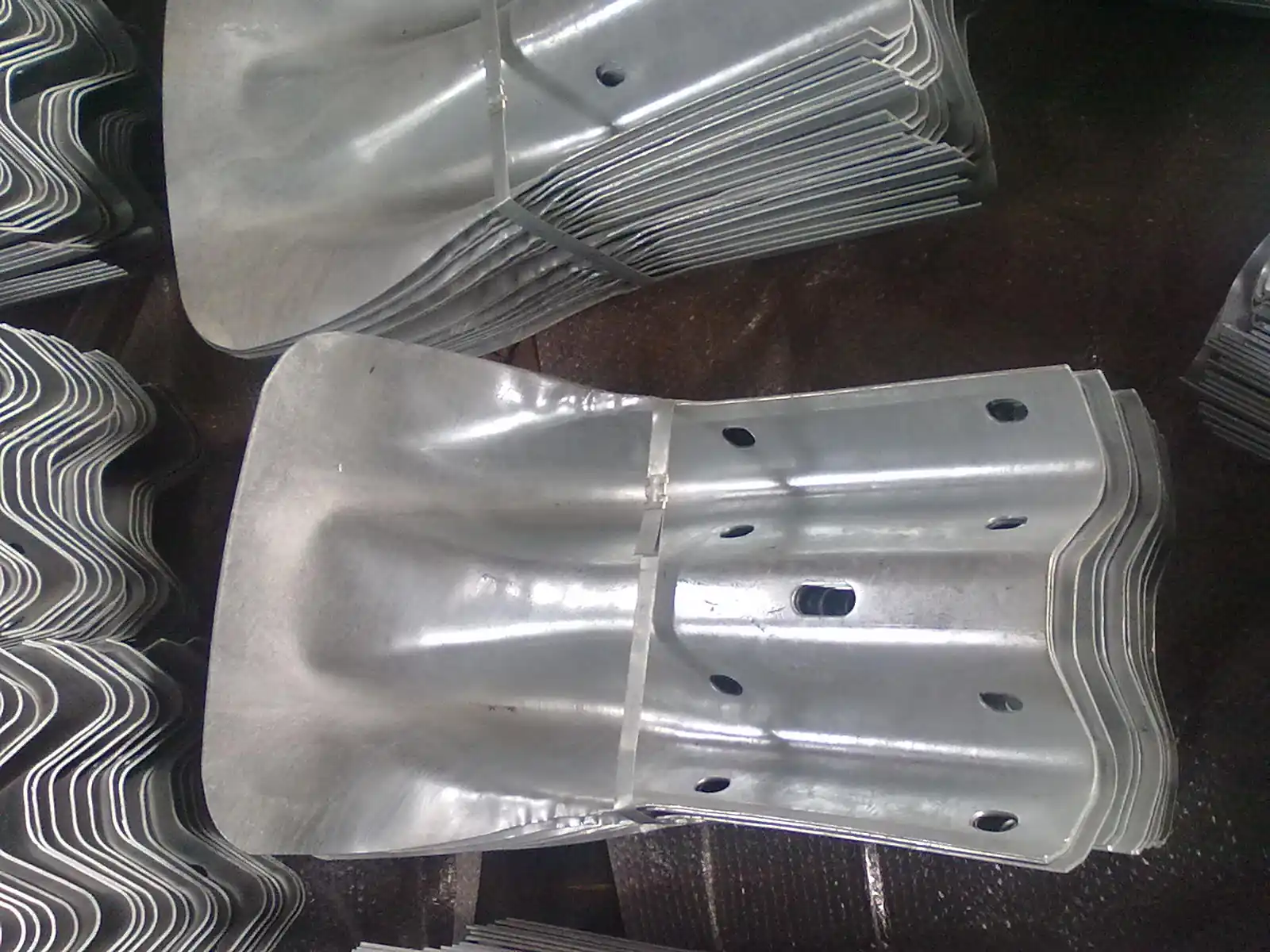 Stacks of galvanized corrugated steel guardrail end components with drilled holes on a