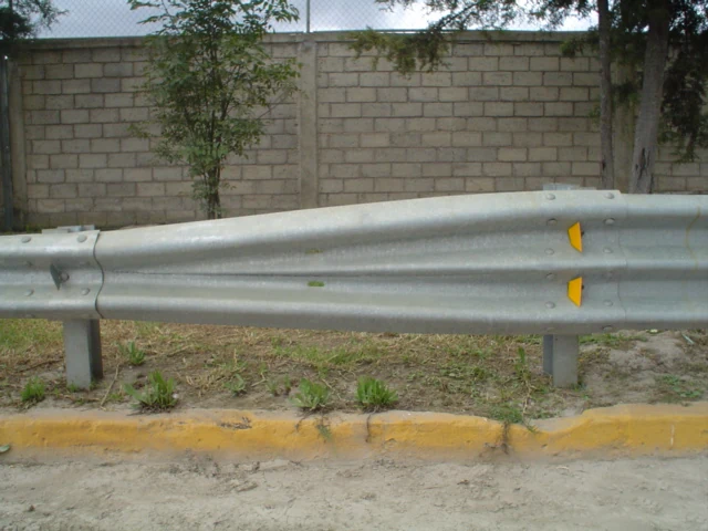 Galvanized W-beam guardrail with yellow reflectors beside a dirt road, concrete block wall and trees behind