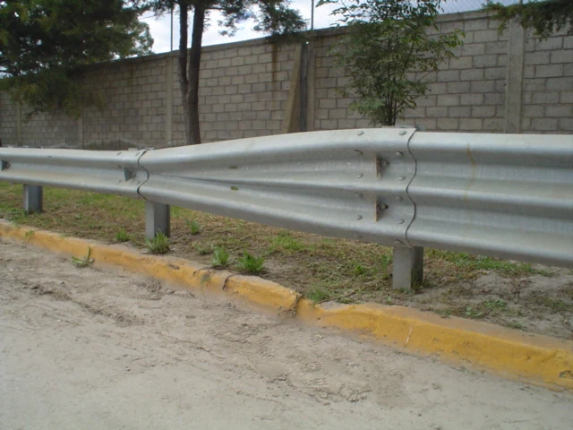Galvanized W-beam highway guardrail on steel posts beside a dirt roadside, with concrete block wall and trees behind