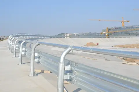 Galvanized W-beam guardrail curving along a concrete bridge, construction cranes visible in the background