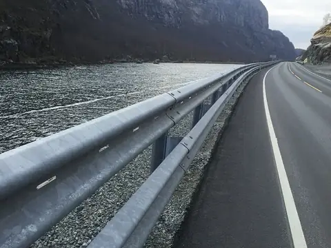 A winding asphalt road alongside a body of water, bordered by a metal guardrail and steep rocky cliffs.