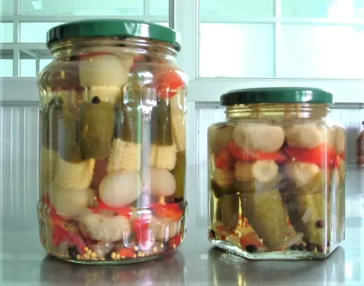 Two glass jars with green lids containing pickled vegetables: baby corn, pearl onions, peppers, cucumbers and