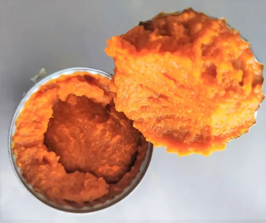 Open tin of smooth orange puree with a scooped round portion lifted out, on a plain light gray surface