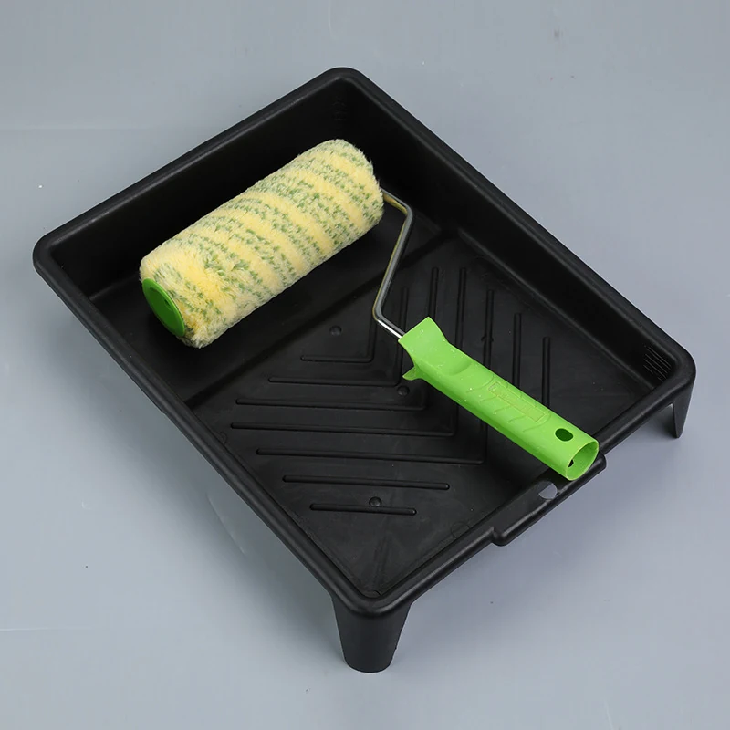 Black plastic paint roller tray with a yellow patterned roller sleeve and green handle on a light gray surface