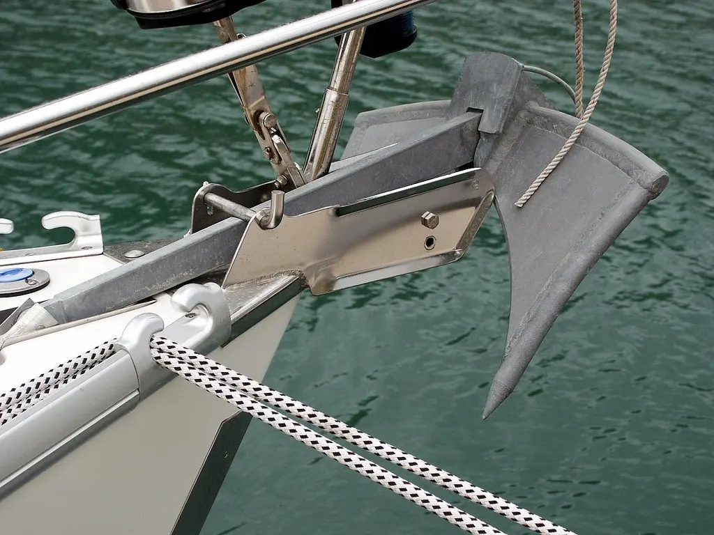 Galvanized steel boat anchor mounted on a bow roller, with a stainless steel railing and green water in the background