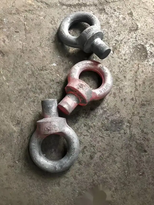 Three forged steel lifting eye bolts, two dark and one with red paint residue, lying on a concrete floor