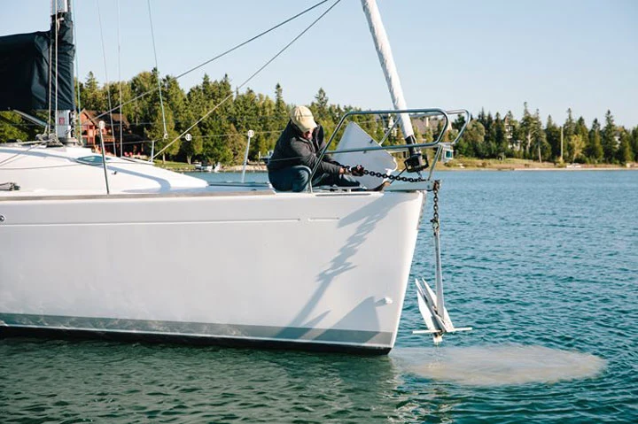 White sailboat at anchor with anchor chain hanging from the bow into calm water, treed shoreline behind