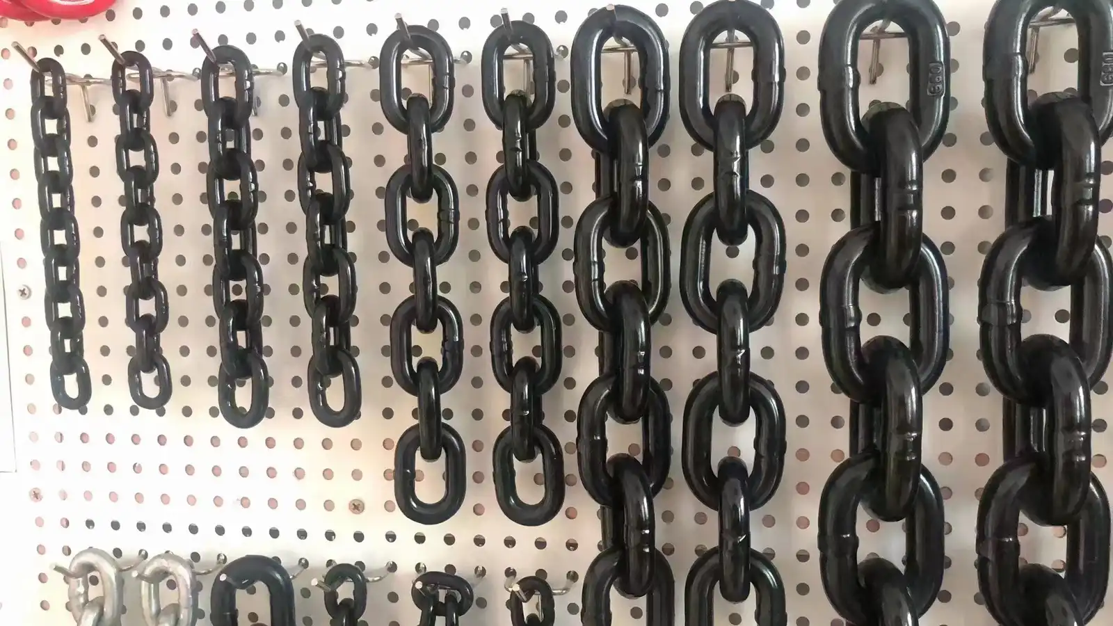 Black lifting chains of various sizes hanging on a white perforated pegboard display, hardware hooks below