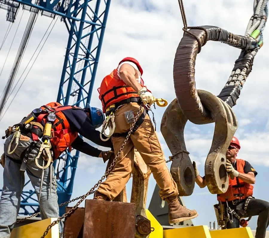 Workers in orange vests and hard hats rigging a large hook near a blue crane
