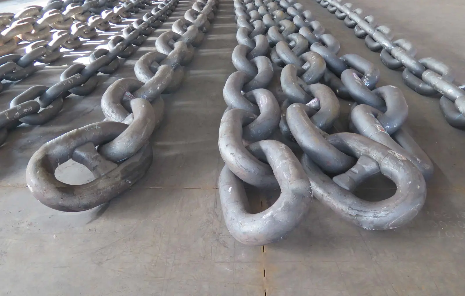 Rows of heavy steel chains with large open links laid out on a bare concrete factory floor