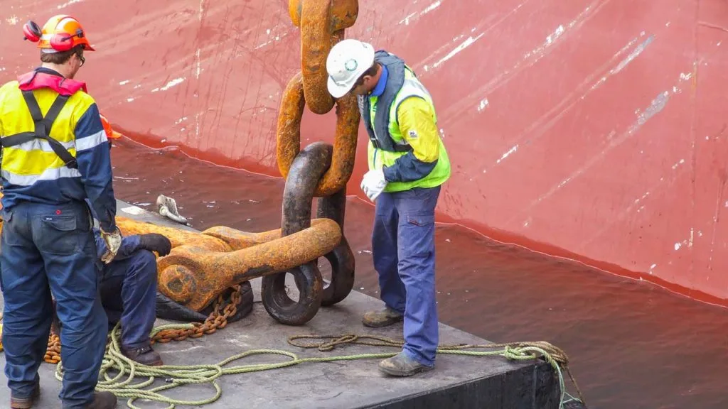 Two workers in hard hats and high visibility vests inspecting a large rusty anchor chain