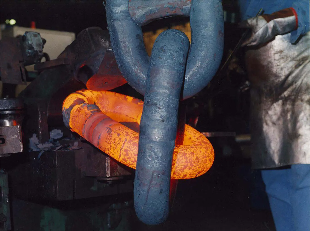 A worker in protective gear uses a large blue hook to manipulate a glowing hot orange metal ring