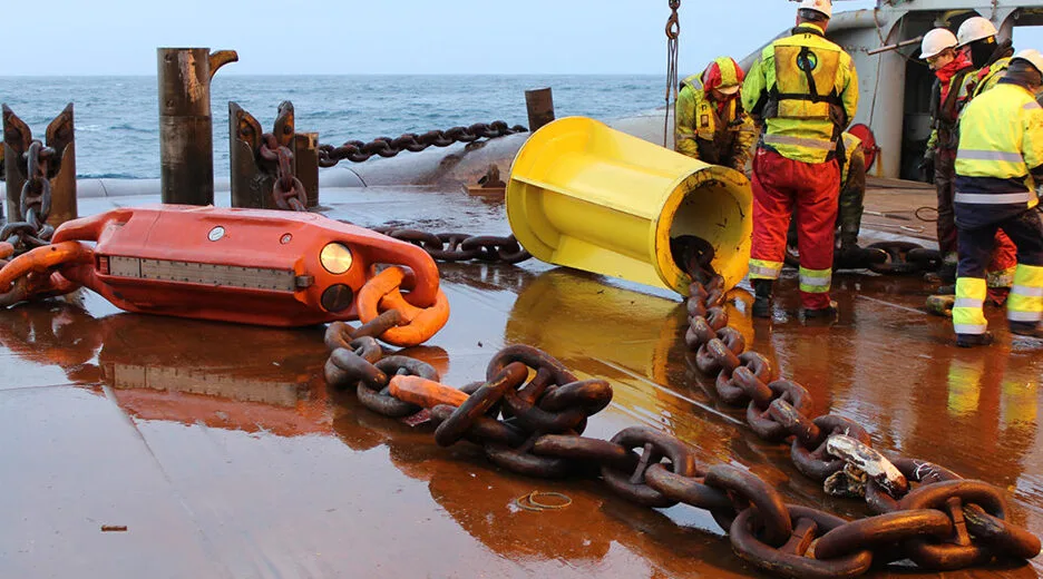 Workers in high visibility gear on a ship deck with heavy anchor chains and a yellow bucket