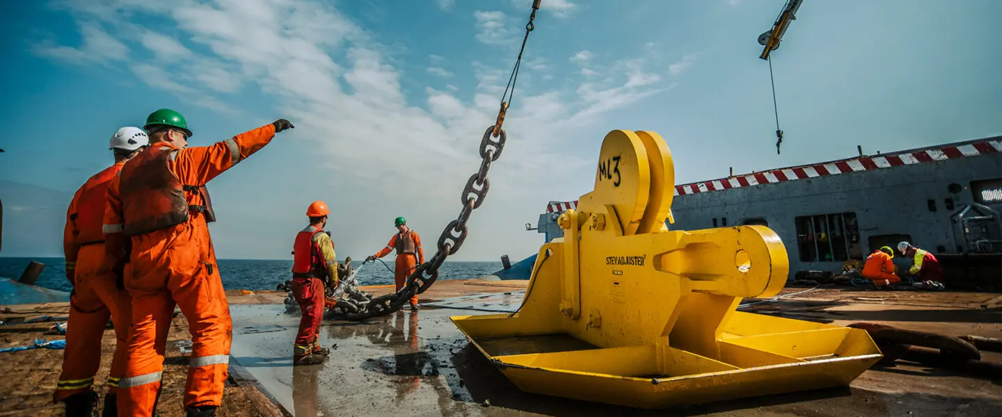 Workers in orange coveralls and hard hats guide a large yellow industrial lifting beam suspended by chains outdoors