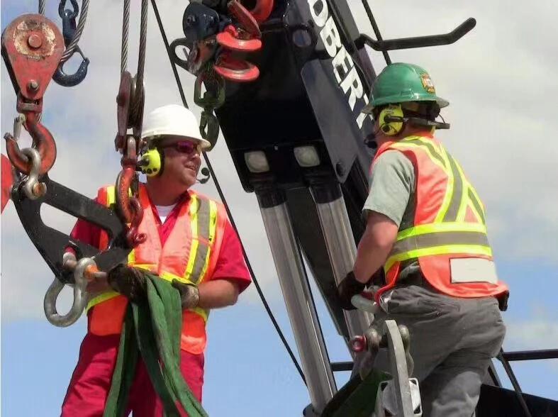 Two industrial workers in hard hats and safety vests operating a crane hook assembly
